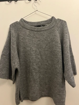 Grå stickad tröja med halvlånga ärmar - Mysig grå stickad tröja med bred ribbad halsringning och halvlånga ärmar. Tröjan är over size och är tillverkad i mjukt material . Enkel och stilren design som passar till det mesta.