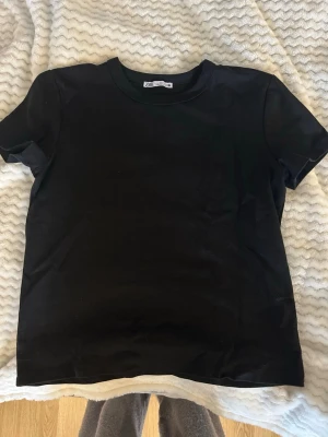 Svart basic t-shirt från Zara - En enkel svart t-shirt från Zara i klassisk modell. T-shirten har rund halsringning och korta ärmar, perfekt till alla typer av outfits. Tillverkad i mjuk bomull för skön känsla hela dagen.