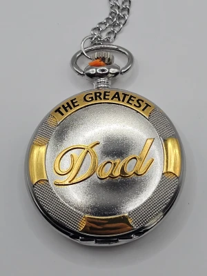 Fickur The Greatest Dad silver/guld "klocka" - Unikt fickur i silver- och guldfärg med texten 'The Greatest Dad' ingraverat på locket. Klockan har en klassisk rund form och kedja i metall, perfekt som personlig accessoar eller present.