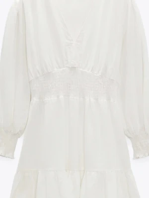 Vit kort klänning med v-ringning - Söt vit kortklänning med v-ringad hals, smockad midja och långa ärmar med resår vid ärmslut. Klänningen har volangdetalj nedtill och är tillverkad i ett lätt tyg som ger en luftig känsla. Perfekt för sommarens alla tillfällen. Den är endast använd 1 gång men sömmen vid v-ringningen har börjat sprättas lite, se bild 4. 