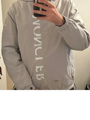 Grå vindjacka från Moncler - Snygg grå vindjacka från Moncler med stor vit logga vertikalt på framsidan. Jackan har dragkedja framtill, två sidofickor med dragkedja och en bröstficka. Elastisk nederkant och ärmslut med knapp. Perfekt för en trendig streetwear-look.
