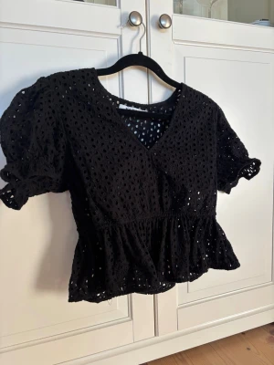 Svart blus med spets och puffärm - Snygg svart blus med v-ringning, puffärmar och volang nedtill. Blusen har ett genombrutet spetsmönster som ger en luftig känsla och feminina detaljer. 