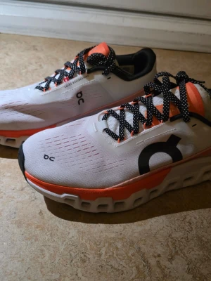 Vita On Running sneakers med orange detaljer - Snygga vita sneakers från On Running med stora svarta loggor på sidan, orange detaljer och svartvitmönstrade skosnören. Ovandelen är i mesh för bra andningsförmåga och sulan har On's karakteristiska CloudTec-design. Perfekta för dig som gillar sportig stil. SNABB AFFÄR 500KR