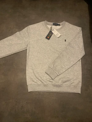 Grå sweatshirt från Polo Ralph Lauren - Klassisk ljusgrå sweatshirt från Polo Ralph Lauren med rund halsringning och diskret broderad logga på bröstet. Tillverkad i mjuk bomullsblandning och har ribbade muddar vid ärmslut och nederkant. Perfekt för en clean och avslappnad stil.
