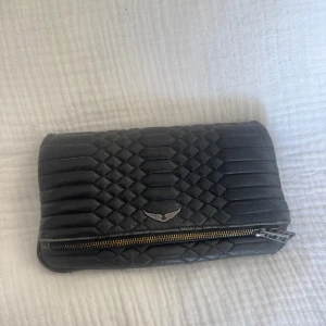 Svart skinn handväska med dragkedja från Zadig - Snygg svart handväska med quiltat mönster och silverfärgade detaljer. Väskan har en dekorativ metallogo framtill och dragkedja både på framsidan och baksidan. Perfekt storlek för det viktigaste och ger en cool vibe till din outfit.