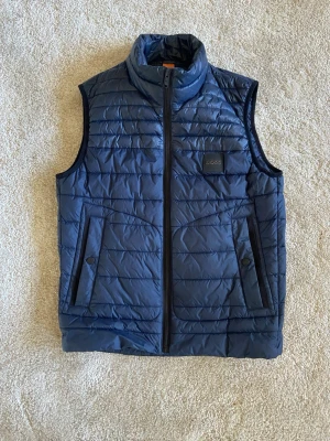 Mörkblå väst från BOSS - Snygg mörkblå väst från BOSS. Västen har två fickor med knappstängning och en innerficka med orange detalj. Perfekt för lager på lager och stilren look.
