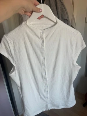 Vit ribbad topp med knappar ☁️☁️ - Säljer en vit ribbad topp med korta ärmar och knappar längs hela framsidan. Toppen har en enkel och clean look, perfekt att styla med jeans eller kjol. Materialet känns mjukt och stretchigt, vilket gör den bekväm att bära.