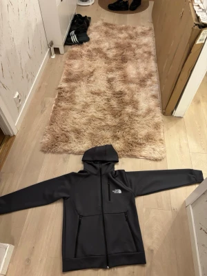 Svart hoodie från The North Face - Snygg svart hoodie från The North Face med dragkedja framtill och två fickor med dragkedja. Klassisk logga på bröstet och skön huva. Materialet känns slitstarkt och passar perfekt för en chill och sportig stil.