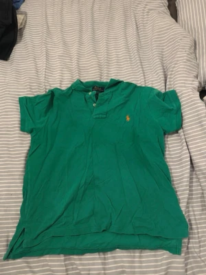 Grön pikétröja från Polo Ralph Lauren - Klassisk grön pikétröja från Polo Ralph Lauren med korta ärmar och broderade loggan i orange på bröstet. Tröjan har krage och knappar vid halsen.