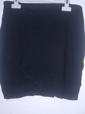 Svart kortkjol från Gina Tricot - Svart kortkjol från Gina Tricot i enkel och stilren design. Kjolen har en omlott-effekt framtill och är tillverkad i ett mjukt material som passar perfekt till olika outfits. Snygg och lätt att matcha med både t-shirts och blusar.