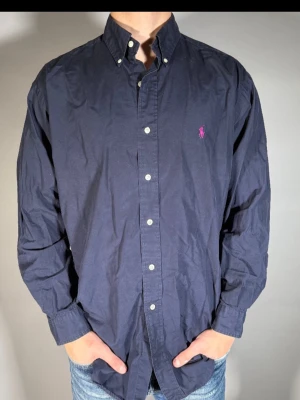 Mörkblå skjorta från Ralph Lauren - Klassisk mörkblå skjorta från Ralph Lauren med button-down krage och lång ärm. Skjortan har vita knappar och den ikoniska rosa broderade loggan på bröstet. Tillverkad i bomull för en skön och avslappnad passform.