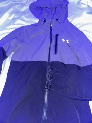 Under Armour vindjacka - Snygg vindjacka från Under Armour i tvåfärgad design, mörkblå och ljusare blå upptill. Jackan har huva, hel dragkedja, bröstficka med dragkedja och logga på bröstet. Tillverkad i lätt och tåligt material, perfekt för blåsiga dagar.