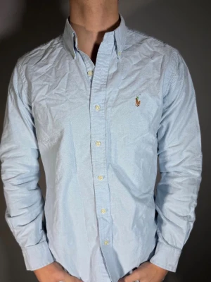 Ljusblå skjorta från Ralph Lauren - Klassisk ljusblå skjorta från Ralph Lauren med button-down krage och broderad logga på bröstet. Skjortan har långa ärmar, knäppning framtill och är tillverkad i bomull. Perfekt för en stilren och avslappnad look.