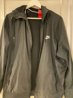 Svart Nike vindjacka med huva - Snygg svart vindjacka från Nike med vit logga på bröstet. Jackan har dragkedja framtill, två fickor med dragkedja och justerbar huva med snören. Perfekt för blåsiga dagar och enkel att matcha med olika outfits.