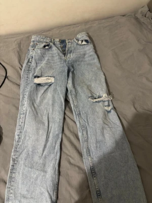 Ljusblå ripped mom jeans - Säljer ett par ljusblå mom jeans med slitna detaljer och hål på båda benen. Jeansen har hög midja, knappgylf och klassisk femficksdesign. Perfekta för en avslappnad och trendig look.