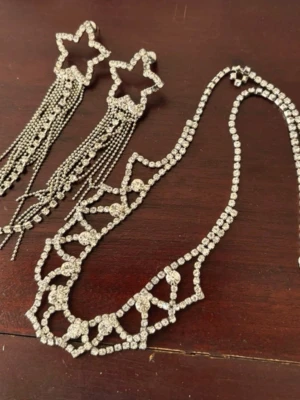 Glittrigt halsband med stjärndetaljer - Säljer ett glittrigt halsband med gnistrande stenar och unika stjärnformade detaljer. Halsbandet har en silverfärgad bas och är dekorerat med små, klara stenar som ger en lyxig känsla. 1 par örhängen med detalj stjärna. Perfekt för dig som vill sticka ut med ett statement-smycke.