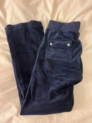 Mörkblå velour mjukisbyxor Juicy Couture - Mörkblå mjukisbyxor i mjuk velour från Juicy Couture. Byxorna har bred resår i midjan med dragsko, två framfickor och en bakficka med tryckknappar. Perfekta för en avslappnad och trendig look.