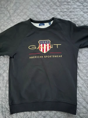 Svart sweatshirt från Gant - Svart sweatshirt från Gant med broderad logga och texten 'American Sportswear' framtill. Tröjan har rund halsringning, ribbade muddar och klassisk passform. Perfekt för en sportig och stilren look. Tyvärr lite sönder vid armbågen men inget som visas jättemycket när man har den på sig.