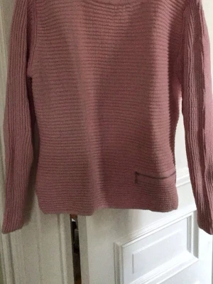 Rosa stickad tröja från Skovhuus - Mysig rosa stickad tröja från Skovhuus med rund halsringning och diskret strukturmönster. Tröjan har långa ärmar och en liten fickdetalj framtill. Perfekt för dig som gillar en enkel men snygg look.