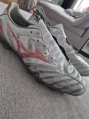 Vita Mizuno fotbollsskor med dobbar - Säljer ett par vita fotbollsskor från Mizuno med röda detaljer och klassisk snörning. Skorna har fasta plastdobbar och är tillverkade i syntetmaterial med sydda förstärkningar framtill för extra kontroll. Perfekta för dig som vill ha stilrena och funktionella skor på planen.