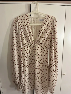 Vit blommig kortklänning från Adoore - Supersöt vit kortklänning från Adoore med små röda blommor, v-ringad hals och långa puffärmar. Klänningen har en feminin passform och är perfekt för vår och sommar. Tillverkad i ett lätt och luftigt material.