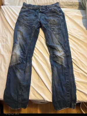 G star jeans 30/32 - Säljer ett par blå jeans med coola patchade detaljer och kontrastsömmar. Jeansen har flera fickor, inklusive en extra ficka på låret, och en avslappnad passform. Materialet är klassisk denim i bomull. Perfekta för en avslappnad och street-inspirerad stil.