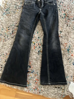 Svarta bootcut jeans med kontrastsömmar - Säljer ett par svarta bootcut jeans med vita kontrastsömmar och klassisk femficksmodell. Jeansen har utsvängda ben och en snygg tvättad look. Perfekta för dig som gillar retrostil och vill ha ett par jeans som sticker ut lite extra.