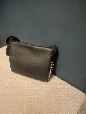 Svart axelväska från Burberry - Snygg svart axelväska från Burberry i läder med klassiskt rutigt mönster i beige, svart, vitt och rött på insidan. Väskan har justerbar axelrem och stilren design med lock. Perfekt för dig som gillar tidlös och trendig stil.