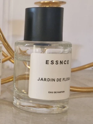 ESSNCE Jardin de Flora parfym - Modern parfymflaska från ESSNCE med doften Jardin de Flora. Flaskan är cylindrisk i genomskinligt glas med svart, matt kork och vit etikett med svart text. Eau de Parfum-variant som ger en exklusiv känsla.