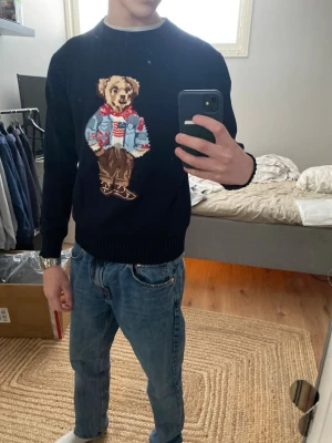 Mörkblå stickad tröja med björnmotiv - Säljer en mörkblå stickad tröja från Ralph Lauren med ikoniskt björnmotiv på bröstet. Björnen har på sig en blå jeansjacka, rödvit tröja och bruna byxor. Tröjan har rund halsringning och ribbade muddar. Perfekt för dig som vill ha en unik och trendig look.