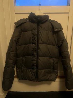 Grå pufferjacka Calvin Klein Jeans - Säljer en grå pufferjacka från Calvin Klein Jeans med hög krage, huva och dragkedja framtill. Jackan har stora vadderade paneler och logotypen tryckt längs dragkedjan. Perfekt för kalla vinterdagar och riktigt snygg streetstil.