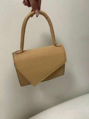 Beige handväska med krokopräglat mönster - Trendig beige handväska i konstläder med krokopräglat mönster och unik triangulär lockdesign. Väskan har ett vadderat handtag som ger en cool och modern look. Perfekt accessoar för att lyfta din outfit.