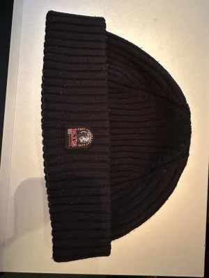 Svart ribbstickad mössa Parajumpers - Svart ribbstickad mössa äkta Parajumpers med uppvikt kant och broderad patch framtill med logga och texten 'P.J.S Anchorage'. Klassisk och stilren modell som håller dig varm under kalla dagar.