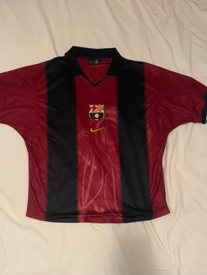 Barcelona x Cactus Jack - Säljer en Barcelona fotbollströja från Nike i röd och svart med breda vertikala ränder. Tröjan har klubbmärket på bröstet, gul Nike-logga och texten 'Cactus Jack', nummer 2 och 'BARÇA' på ryggen. Klassisk krage och korta ärmar i lätt, ventilerande material.