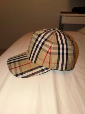 Burberry klassisk rutig keps - ((Möts bara inom gbg, vid snabb affär kan priset sänkas)) Säljer en snygg keps från Burberry med det ikoniska beige, svarta, vita och röda rutmönstret. Kepsen har justerbar snapback och en svart patch med texten 'BURBERRY LONDON ENGLAND' bak. Perfekt accessoar för en trendig streetwear-look.
