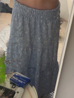 Blå blommig långkjol med volang - Söt blå långkjol med små vita blommor och volangdetaljer. Kjolen har bred resår i midjan för skön passform och ett luftigt fall. Perfekt för dig som gillar en romantisk och avslappnad stil. säljer den då den är för lång för mig och inte kommer till användning, skriv om ni har frågor