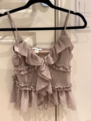 Ljusbeige volang topp  - Supersöt ljusbeige linnetopp med volanger och smala axelband. Toppen har ribbad struktur, omlottdetalj framtill och flera lager volanger. Använd 2-3 gånger💖