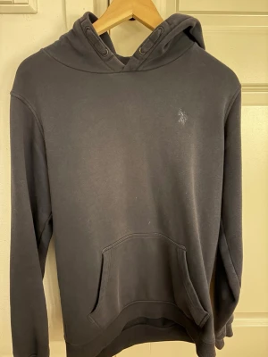Navy hoodie från U.S. Polo Assn. - Snygg mörkgrå hoodie från U.S. Polo Assn. med broderad logga på bröstet. Klassisk känguruficka framtill och justerbar huva med dragsko. Tillverkad i mjukt och bekvämt material, perfekt för chill dagar eller när du vill ha en clean look. Sitter som S/M.