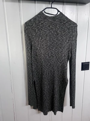 Mörkgrå stickad polotröja - Mysig mörkgrå stickad lite längre polotröja med lång ärm och slits i sidan. Tröjan har en smal passform och är tillverkad i mjukt, melerat material som känns skönt mot huden. Perfekt för lager på lager och enkel att matcha med jeans eller kjol.