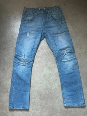 Ljusblå jeans från Jack & Jones - Säljer ett par ljusblå jeans från Jack & Jones med slitningar och förstärkta sömmar vid knäna. Jeansen har flera fickor, raka ben och coola detaljer som ger en streetwear-vibe. Perfekta för dig som gillar en avslappnad men trendig stil.