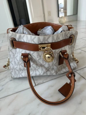 Michael Kors beige handväska - Snygg beige handväska från Michael Kors med MK-logga i mönstret, bruna läderdetaljer och guldfärgade metalldelar. Väskan har dubbla handtag, kedjerem och ett dekorativt hänglås framtill. Tillverkad i tåligt material med vattenavvisande yta.