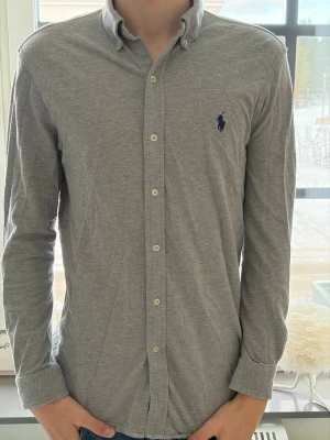 Grå skjorta från Ralph Lauren - Klassisk grå skjorta från Ralph Lauren med lång ärm och knappar framtill. Skjortan har button-down krage och den ikoniska mörkblå loggan broderad på bröstet. Tillverkad i mjuk bomull för en bekväm känsla. Priset är inte hugget i sten!💎✅
