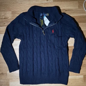 Zip-Knit Polo Ralph Lauren - Zip-Knit från Polo Ralph Lauren med halv dragkedja och röd broderad logga på bröstet. Tröjan har hög krage och långa ärmar, perfekt för kyligare dagar. Klassisk och tidlös design som passar till det mesta.TVEKA INTE ATT SKICKA EN BUD:)