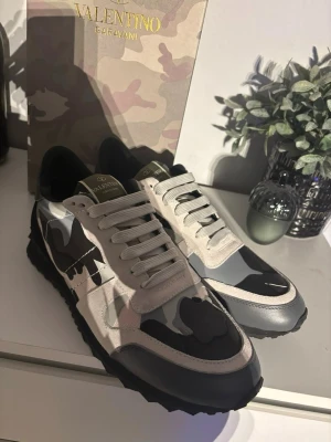 Valentino Garavani camo sneakers - Snygga sneakers från Valentino Garavani med svart, grå och vit camo-mönster. Skorna har vita snören, svarta detaljer och en robust sula. Materialmix av mocka, läder och textil ger en exklusiv känsla. Perfekta för dig som vill sticka ut med stil.