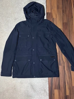 Mörkblå vårjacka från Peak Performance - Snygg mörkblå jacka från Peak Performance med fyra stora fickor framtill och justerbar huva. Jackan har en stilren design med tryckknappar och diskret logga på ena fickan. Perfekt för kalla dagar och enkel att matcha med olika outfits.