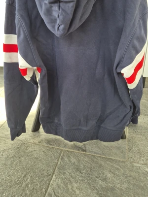 Blå hoodie från Gant med röd/vit detalj - Mörkblå hoodie från Gant med huva och dragsnören. Framtill finns ett stort Gant-tryck och en sköld med stjärnor och ränder. Tvärgående vita och röda ränder över bröstet och armarna. Tillverkad i mjukt bomullsmaterial, perfekt för en avslappnad stil.