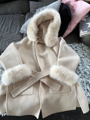Beige kappa med päls och huva Giovanni Paris - Snygg beige kappa med stor huva och breda pälsdetaljer vid huva och ärmslut. Kappan har knytskärp i midjan och två fickor framtill. Perfekt för dig som vill ha en mysig och trendig look under kyliga dagar.