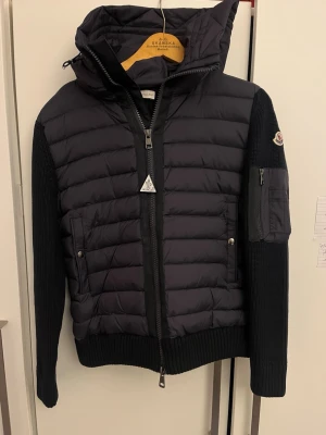 Svart hybrid cardigan från Moncler - Snygg cardigan jacka från Moncler med vadderad framsida och huva. Ärmar och rygg är stickade i mjukt material, medan framsidan är quiltad. Dragkedja framtill, fickor med tryckknappar och en Moncler-logga på ärmen. Perfekt för lager på lager och streetstyle.