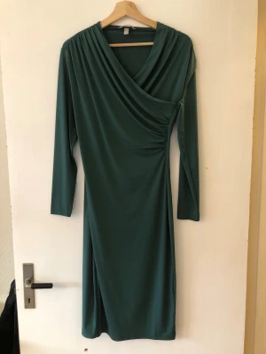 Grön omlottklänning med lång ärm - Stilren grön klänning med omlott-design och rynkade detaljer vid sidan. Klänningen har lång ärm och v-ringad hals, vilket ger en elegant och modern look. Perfekt för dig som gillar enkel men snygg stil. Polyester