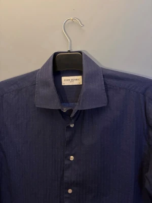 Mörkblå skjorta från John Henric - Stilren mörkblå skjorta från John Henric med diskret randigt mönster och klassisk krage. Skjortan har vita knappar och lång ärm, perfekt för en clean och snygg look. Tillverkad i bomull för skön känsla.